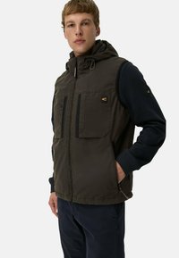 Kapuzensleeveless-Weste in Olivgrün, ausgestattet mit zwei Vorder- Taschen, einem durchgehenden Reißverschluss und verstellbaren Kordeln, kombiniert mit einem navyfarbenen Langarmshirt.