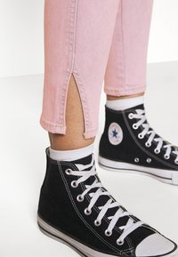 Jeans de mezclilla rosa con una abertura en la parte inferior, combinados con zapatillas altas negras con cordones blancos y un logotipo en el tobillo.