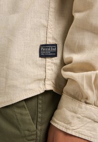 Beige linnen shirt met opgerolde mouwen, met een marineblauw merkembleem. Olijfgroene corduroy broek is gedeeltelijk zichtbaar aan de onderkant.