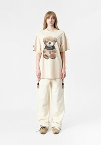 Camiseta oversized beige con un gráfico de un oso de peluche, combinada con pantalones sueltos color blanco roto. Los zapatos son marrones con detalles blancos y cordones.