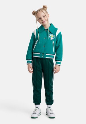 Κορίτσι με διπλούς κότσους που φοράει μπουφάν τένις σε τυρκουάζ και λευκό, ταιριαστά παντελόνια, λευκές κάλτσες και λευκά αθλητικά παπούτσια με πράσινες λεπτομέρειες.