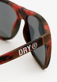 Lunettes de soleil en plastique avec une monture à motifs rouge et marron, verres foncés, et un logo "DRY®" blanc sur la branche.