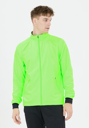 Junger Mann trägt eine leuchtend grüne Jacke mit Reißverschluss und schwarze Hose, blickt nach rechts vor schlichtem weißem Hintergrund.