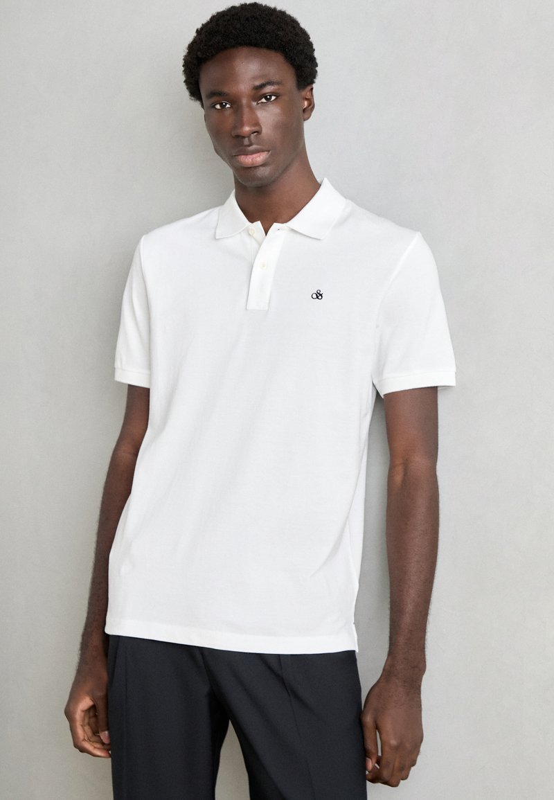 Hvit polo skjorte laget av bomull, med krage, korte ermer og en liten svart logo på brystet, sammen med mørke bukser.