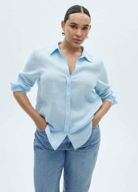 Camicia azzurro chiaro con bottoni in un tessuto leggero, caratterizzata da maniche lunghe arrotolate all'altezza dei gomiti e una vestibilità rilassata. Abbinata a jeans blu.