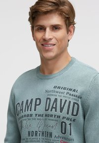Hellblauer Strickpullover mit Rundhalsausschnitt und Schriftzug "CAMP DAVID" in fetter schwarzer Schrift. Der Text enthält geografische Angaben.