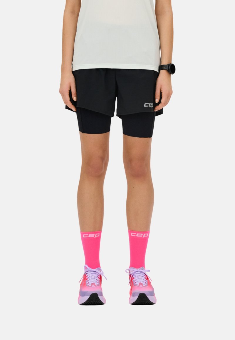 Person trägt schwarze, mehrlagige Laufshorts, leuchtend pinke, knielange Socken mit dem Schriftzug „cep“, pink-lila Laufschuhe und ein weißes Oberteil.