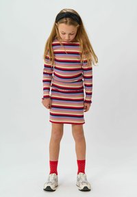 Rihmade pika varrukaga topp ja sobiv mini-seelik erinevates värvides. Tekstureeritud materjal, millel on volangilised aksessuaarid ja vormiv siluett. Punase täpikaga sokid ja sneakersid.