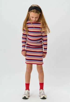 Rihmade pika varrukaga topp ja sobiv mini-seelik erinevates värvides. Tekstureeritud materjal, millel on volangilised aksessuaarid ja vormiv siluett. Punase täpikaga sokid ja sneakersid.