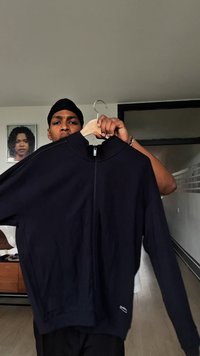 Osoba koja nosi crni durag drži crnu jaknu na zatvaranje na drvenom vješalici unutra, s okvirom portreta u pozadini.