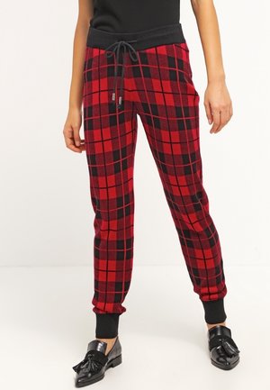 Kobieta nosząca czerwono-czarne spodnie w kratę ze ściągaczami w pasie i czarnymi ściągaczami u dołu nogawek, w połączeniu z czarnymi mokasynami z frędzlami, stojąca na białym tle.