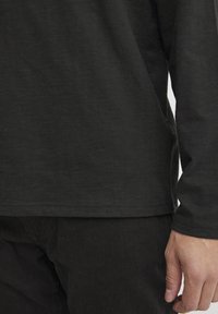 Long-sleeve black shirt, soft texture, round neckline, and side slits at the hem. Detail of hand resting on black fabric below.