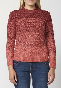 Knit sweater with a high neck and mixed shades of red, featuring a textured pattern and ribbed cuffs and hem. Paired with denim jeans.