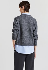 Grauer gestrickter Pullover mit ribbeligem Saum und Bündchen, getragen über einem hellblauen Hemd mit Kragen, das eine entspannte Passform und Textur zeigt.