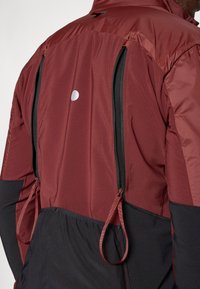 Giacca bordeaux e nera con cerniere, materiali testurizzati e un punto riflettente sulla parte alta della schiena; include dettagli arricciati e design a pannelli.