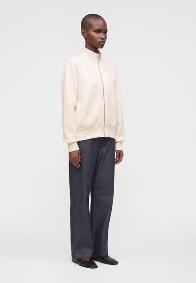 Personne debout portant une veste crème zippée, un pantalon large foncé et des chaussures noires, sur un fond clair uni, légèrement tournée vers la droite.