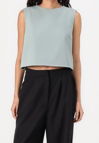 Donna con top azzurro chiaro senza maniche e pantaloni neri larghi a vita alta su sfondo bianco.