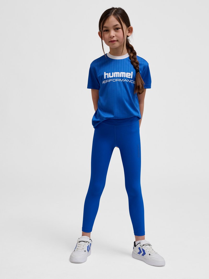 Dziewczyna z długim warkoczem, ubrana w niebieski t-shirt "hummel PERFORMANCE", pasujące niebieskie legginsy oraz białe buty sportowe z niebieskimi akcentami, stoi na szarym tle.