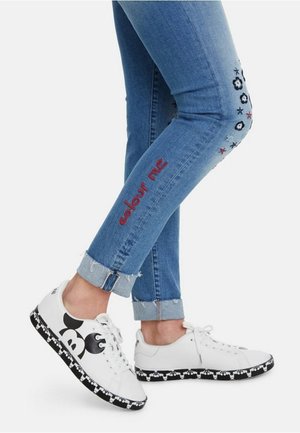 Jean bleu avec revers, brodé de fleurs et le texte "colour me", porté avec des baskets blanches ornées de motifs noirs de Mickey Mouse.