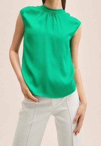 Haut sans manches vert avec un col froncé, en tissu lisse, associé à un pantalon blanc. Coupe simple et arrondie à l'ourlet.