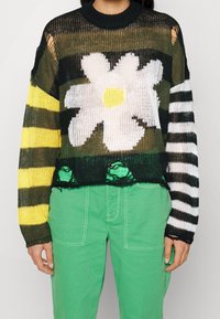Pull court en tricot avec un grand motif de marguerite blanche, des rayures vertes et noires, et des accents jaunes ; bords usés et épaules dégagées.