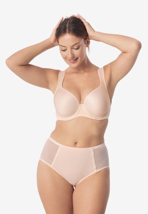 Kobieta w jasnobeżowym biustonoszu i figach z wysokim stanem, stojąca z rękami na głowie, uśmiechająca się, na jednolitym białym tle.