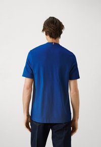 Modré tričko s krátkým rukávem, vyrobené z měkké látky, má jemnou texturu a malý červenobílý detail na výstřihu.