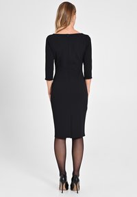 Schwarzes, tailliertes Kleid mit drei Viertelärmeln, knielangem Saum, glatter Textur und einem dezente Reißverschlussdetail auf der Rückseite. Getragen mit schwarzen Absätzen.