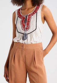 Vit ärmlös topp med röd broderi och svarta detaljer; parat med tanfärgade högmidjade byxor med framveck.