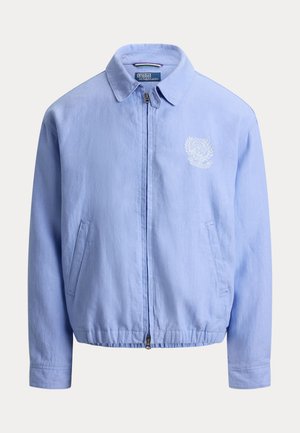 Veste bleu clair à fermeture éclair avec col, ourlet élastique, poches latérales et petit écusson blanc brodé sur la poitrine.