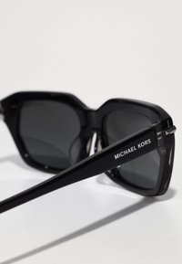 Čierne plastové slnečné okuliare s tmavými sklami, s ostrým dizajnom. Obsahujú logo na bočnej strane: "MICHAEL KORS." Hladký povrch, lesklá úprava.