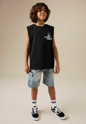 Berniukas stovi rankos šonuose, dėvi juodą berankovį marškinėlį su užrašu „Summer Vibes“, šviesius džinsinius šortus, dryžuotas kojines ir juodus sportbačius.