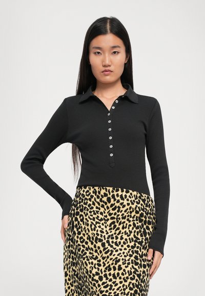 O femeie cu păr lung și întunecat, îmbrăcată cu un top negru cu mâneci lungi și butoni, purtând o fustă bej cu imprimeu leopard negru, pozând cu mâna pe șold.