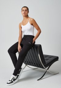 Zwarte joggers met een trekkoord in de taille, een klein geborduurd logo aan de voorkant, gecombineerd met een witte tanktop en zwarte sneakers.