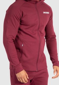 Sudadera con cremallera en burdeos hecha de un tejido suave y texturizado. Presenta un cuello alto, cremallera frontal y un pequeño logo en el pecho. Bolsillos laterales.