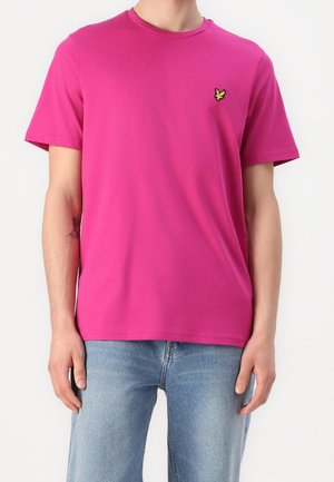 Mies, jolla on kirkkaan pinkki lyhythihainen t-paita, jonka rinnassa on pieni keltainen linnunlogo, yhdistettynä vaaleansinisiin farkkuihin.