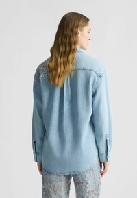 Camicia in denim blu chiaro con maniche lunghe, chiusura frontale con bottoni e dettagli in pizzo sulle spalle. Vestibilità rilassata con orlo curvo.