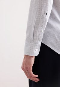 Primo piano del braccio sinistro di una persona che mostra il polsino di una camicia a maniche lunghe grigio chiaro con motivo, con bottoni bianchi e pantaloni neri.
