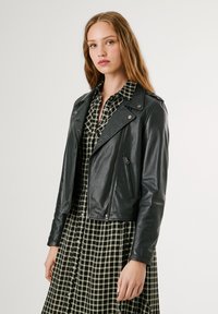 Jaqueta de motociclista de couro preto com fechos éclair e ombreiras, usada sobre um vestido xadrez verde e branco. Textura suave, design ajustado.