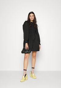 Rochie neagră cu mâneci bufante, bust smocks și închidere cu nasturi. Accesorizată cu o poșetă neagră, tocuri galbene și șosete cu buline.