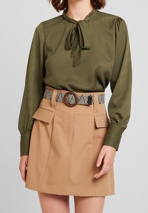 Kvinde iført en olivengrøn bluse med lange ærmer og bindebånd ved halsen, tanfarvet højtaljet nederdel med frontlommer og mønstret bælte med rund spænde.