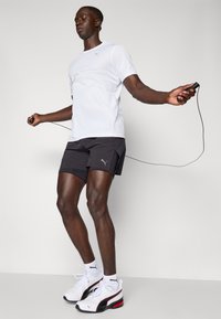 Hvit kortermet treningsskjorte, svarte shorts og hvite joggesko med røde detaljer. Personen hopper tau mot en lys bakgrunn.