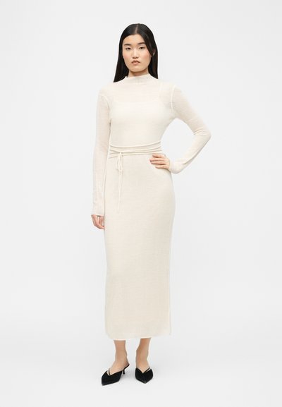 Femme debout, main sur la hanche, portant une robe en maille côtelée crème à manches longues et des escarpins noirs à bouts pointus sur fond blanc.