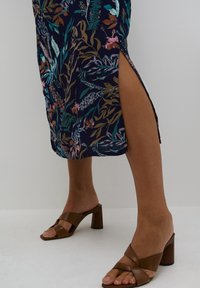 Vestido floral azul marino con aberturas laterales, que presenta patrones botánicos en verde y rosa. Sandalias de tacón cuadrado marrones con diseño de tiras cruzadas.