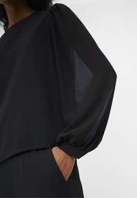 Blouse noire avec manches longues transparentes, encolure ronde et poignets élastiques. Coupe ample avec une texture lisse. Associée à un pantalon noir.