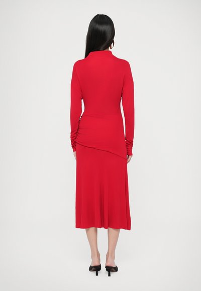 Κόκκινο φόρεμα με μακριά μανίκια και ψηλό λαιμό, με ρίγες στη μέση και φαρδιά φούστα που ρέει. Λείο ύφασμα και μήκος μέχρι τη μέση της γάμπας.