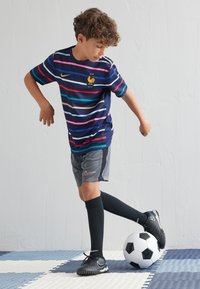 Camiseta de fútbol a rayas marino con líneas multicolores, pantalones cortos grises, calcetas negras y botas, de pie sobre un balón de fútbol blanco y negro.