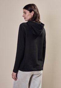 Svart huva tröja med långa ärmar och avslappnad passform, sedd bakifrån, tillsammans med ljusa, strukturerade byxor.