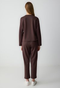 Felpa marrone e pantaloni abbinati realizzati in tessuto morbido; vestibilità comoda con maniche lunghe, scollo a girocollo e vita elastica. Sneakers bianche.
