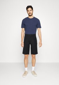Navy T-Shirt mit kurzen Ärmeln, kombiniert mit knielangen schwarzen Shorts. Legere beigefarbene Sneakers und weiße Knöchelsocken vervollständigen das Outfit.